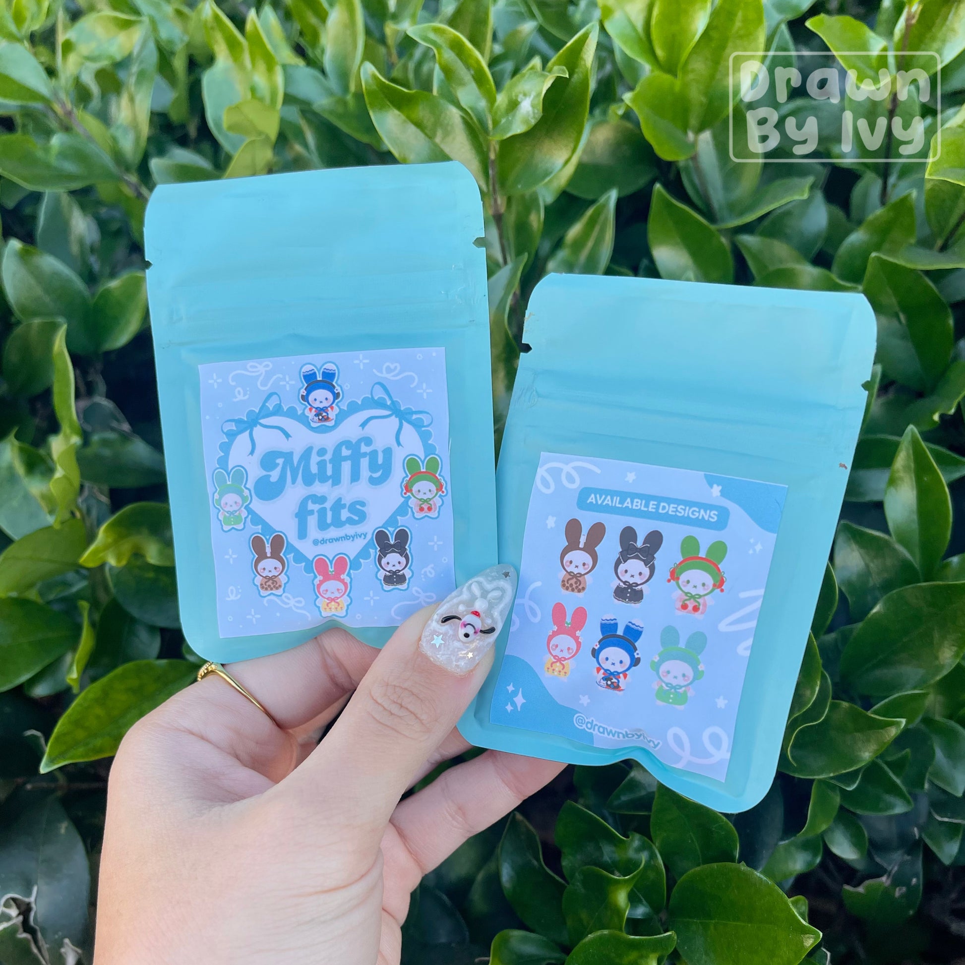 Two blue packages with cartoon characters held in front of green foliage, branded 'Drawn by Ivy'.